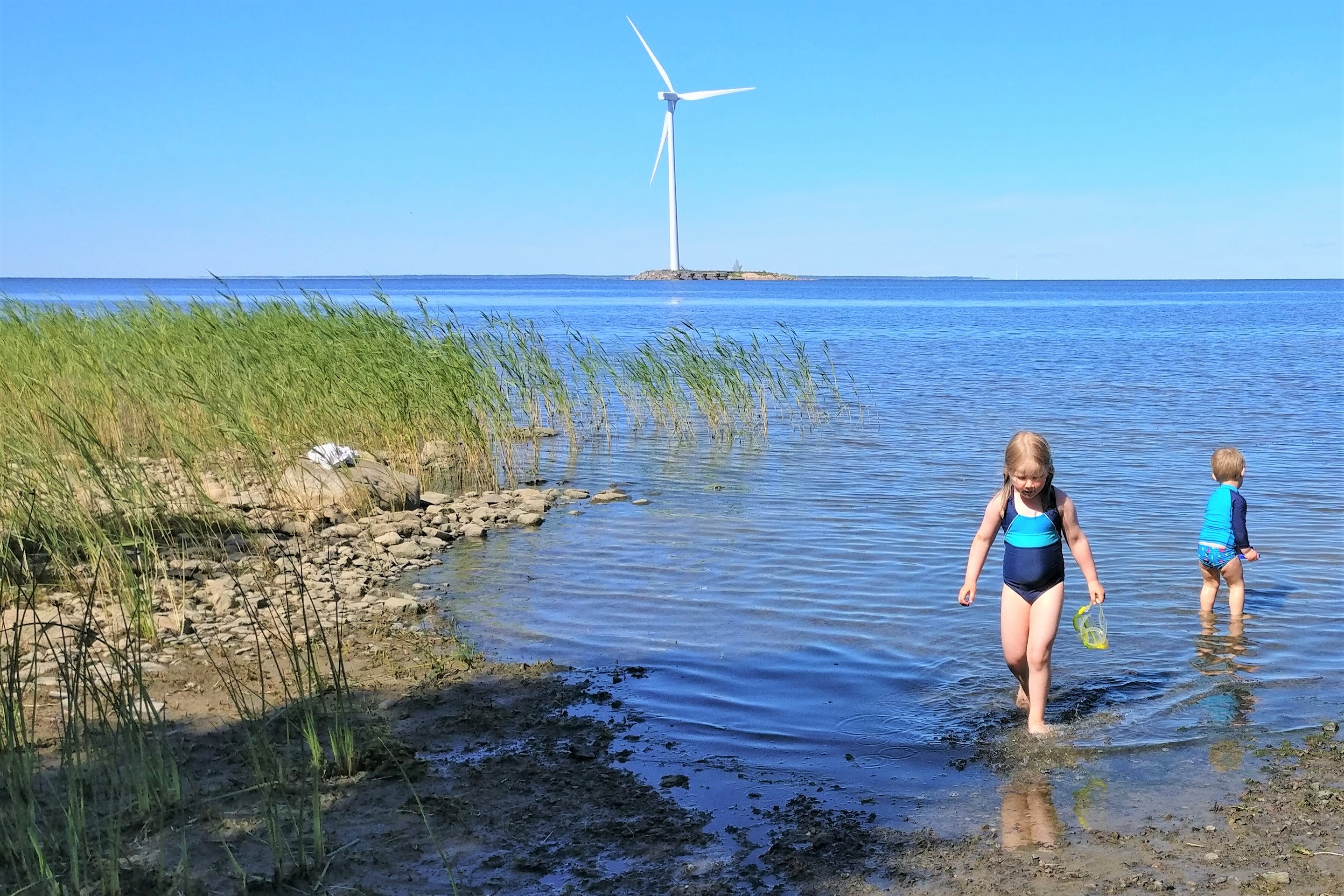 Valokuva, jossa lapset ovat rannassa uimassa ja taustalla näkyy tuulivoimala.